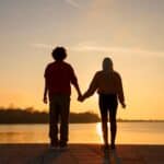 Couple holding hands by a serene lake at sunset, symbolizing support and exploring how to work through trauma in therapy.