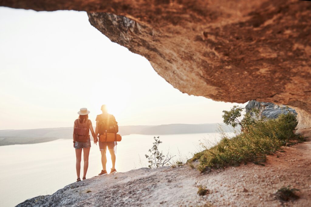 A couple stands with their backs facing a rock and lake view at dawn, sunlight surrounding them as they share a quiet, reflective moment