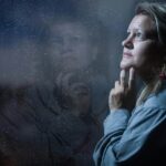 A middle-aged woman gazes out a window covered in rain droplets, reflecting contemplation, emotion, and inner processing.