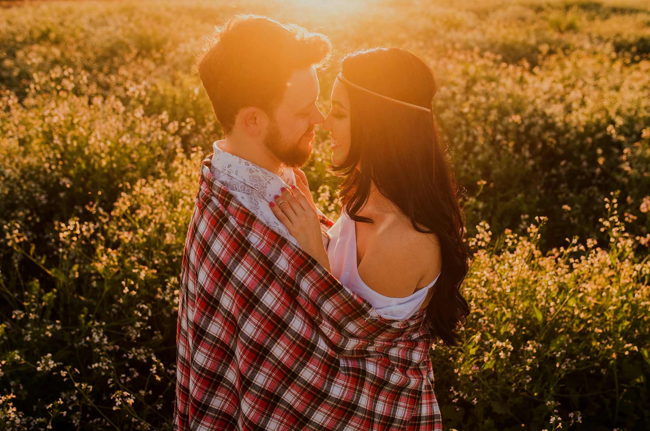Couple standing close in a sunlit field with flowers behind them, intimate moment of connection and warmth