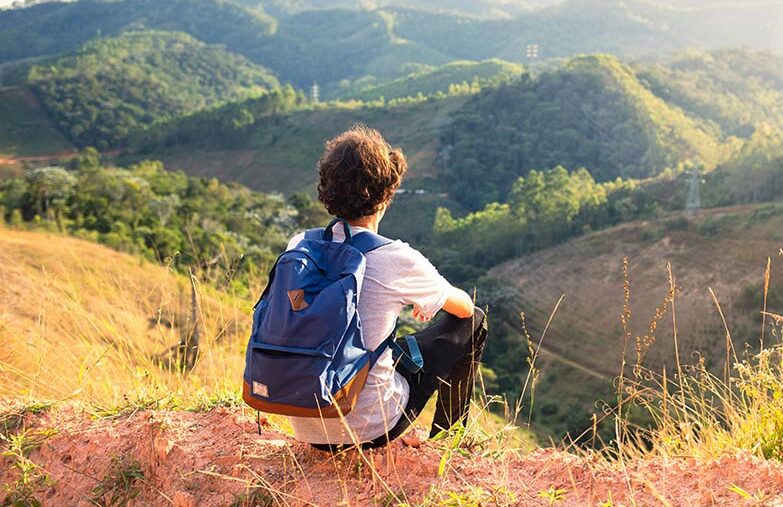 Man seated on a mountaintop facing a wide green mountain landscape, posture relaxed, conveying reflection and presence