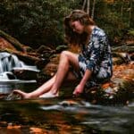  A woman sitting by a stream with feet in the water, looking down peacefully