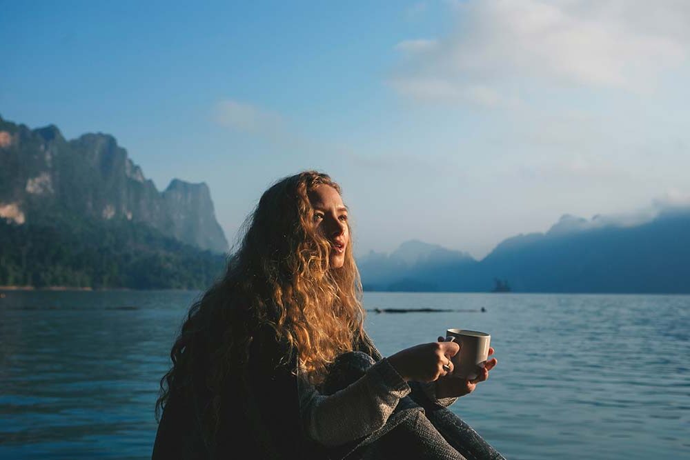 A woman holding coffee overlooking a mountain lake with sunlight on her face, with a peaceful expression.