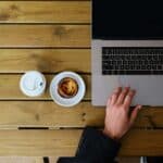 Hands resting near a laptop, coffee, and dessert on a table, casual workspace suggesting focus and comfort