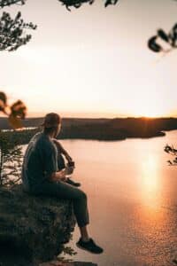  A man sitting on a rock overlooking a lake or river, still and reflective, taking in the quiet and space around him slowly