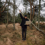  A man standing  in a quiet forest with a laptop surrounded by trees and a natural woodland landscape