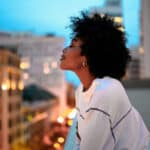 African American woman standing on a balcony with eyes closed, overlooking the city, in a peaceful moment of calm