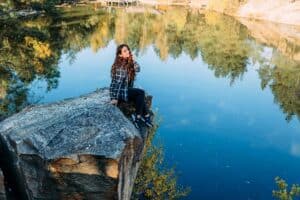  A woman sitting on the edge of a rock by a calm lake, smiling softly while taking in the view and feeling a quiet, peaceful moment