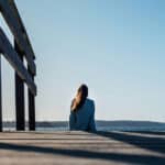 Woman sitting on a wooden deck by a riverbank overlooking mountains, trees, and a calm lake view