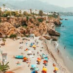 Sunny beach with colorful umbrellas, people relaxing by the ocean, and mountains in the distance