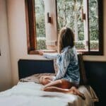  A woman sitting on a bed looking out a window, soft light coming in as she pauses in a quiet moment of thought and reflection