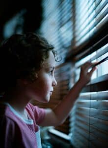 Child looking out through window blinds, soft light casting shadows across their face, capturing quiet reflection and emotion.
