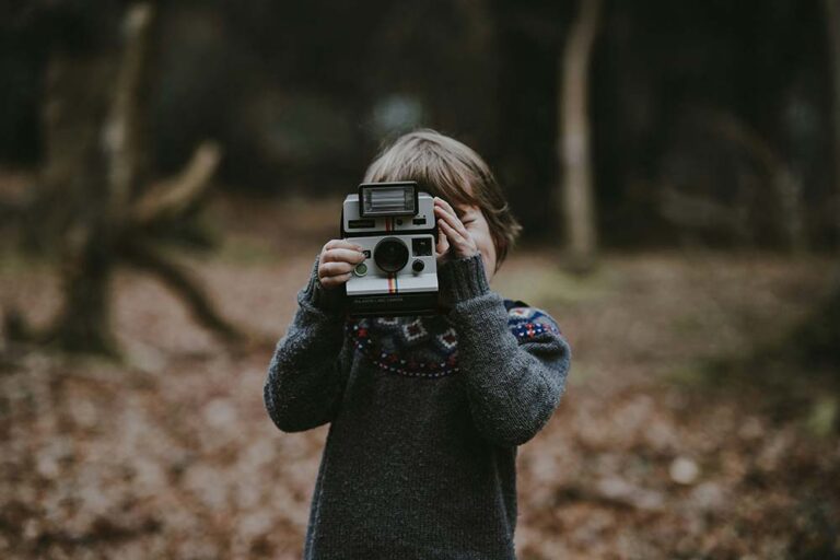 A young child in a grey patterned sweater stands in a wooded area, holding a vintage instant camera up to their face as if taking a photo.