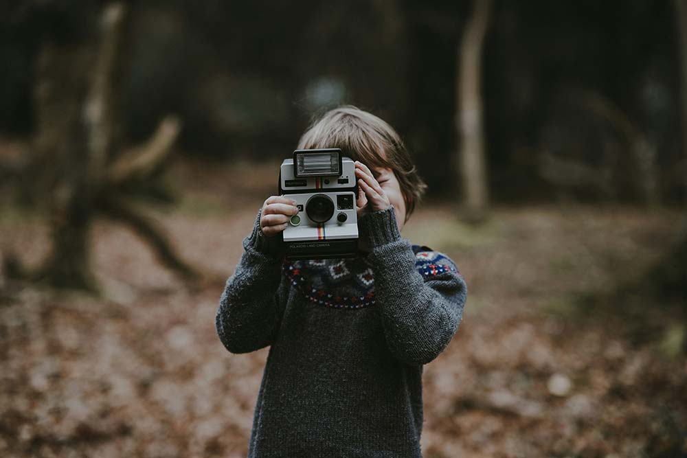 A young child in a grey patterned sweater stands in a wooded area, holding a vintage instant camera up to their face as if taking a photo.