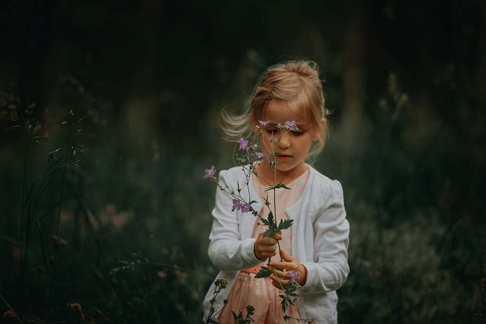 
A young girl with blonde hair stands in a grassy field, wearing a white cardigan over a pink dress.