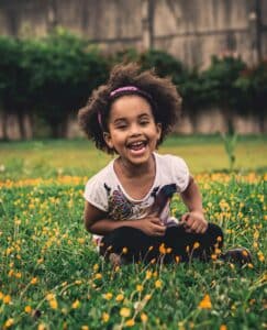  An African American girl sitting in the grass with flowers, smiling and expressing joy, safety, and emotional freedom.