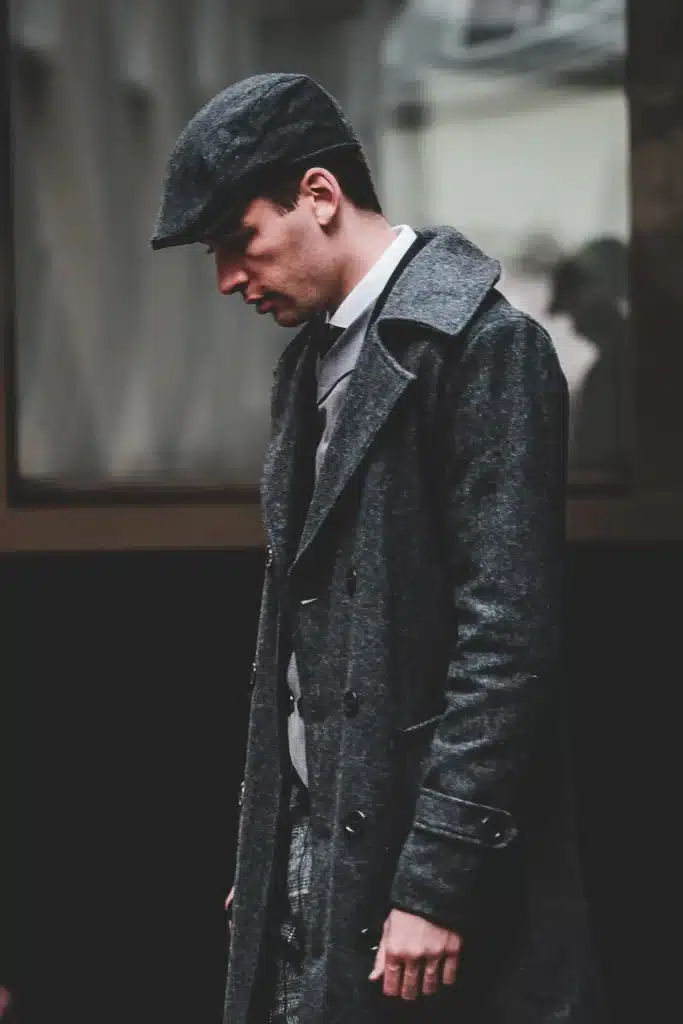 A young man in profile wearing a dark grey flat cap and a matching wool overcoat looking downward.