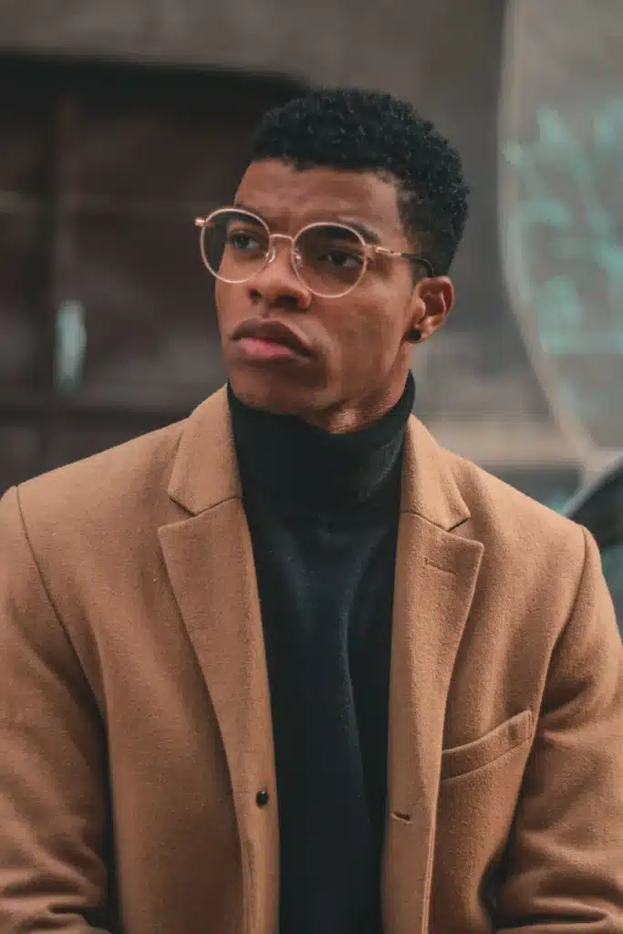A young man wearing round wire-frame glasses, a black turtleneck, and a light brown coat looking off-camera.