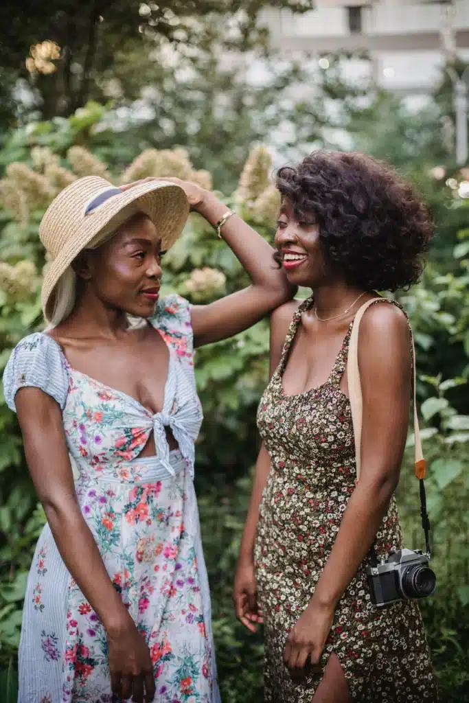 Two women wearing floral sundresses standing close together in a lush park, one touching her straw hat while the other smiles with a vintage camera slung over her shoulder.