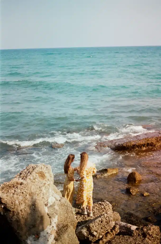 Two women in matching yellow floral dresses standing together on a rocky shore looking out at turquoise ocean waves.