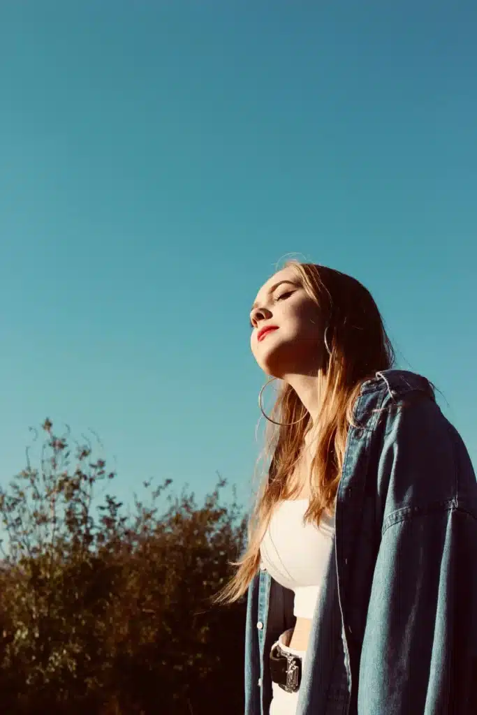 A young woman with her eyes closed facing sunlight, wearing an oversized denim jacket, white top, and hoop earrings against a clear blue sky.