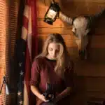 A young woman in a maroon top sitting inside a rustic wooden cabin, holding a vintage camera beneath a hanging lantern and a mounted bull skull.